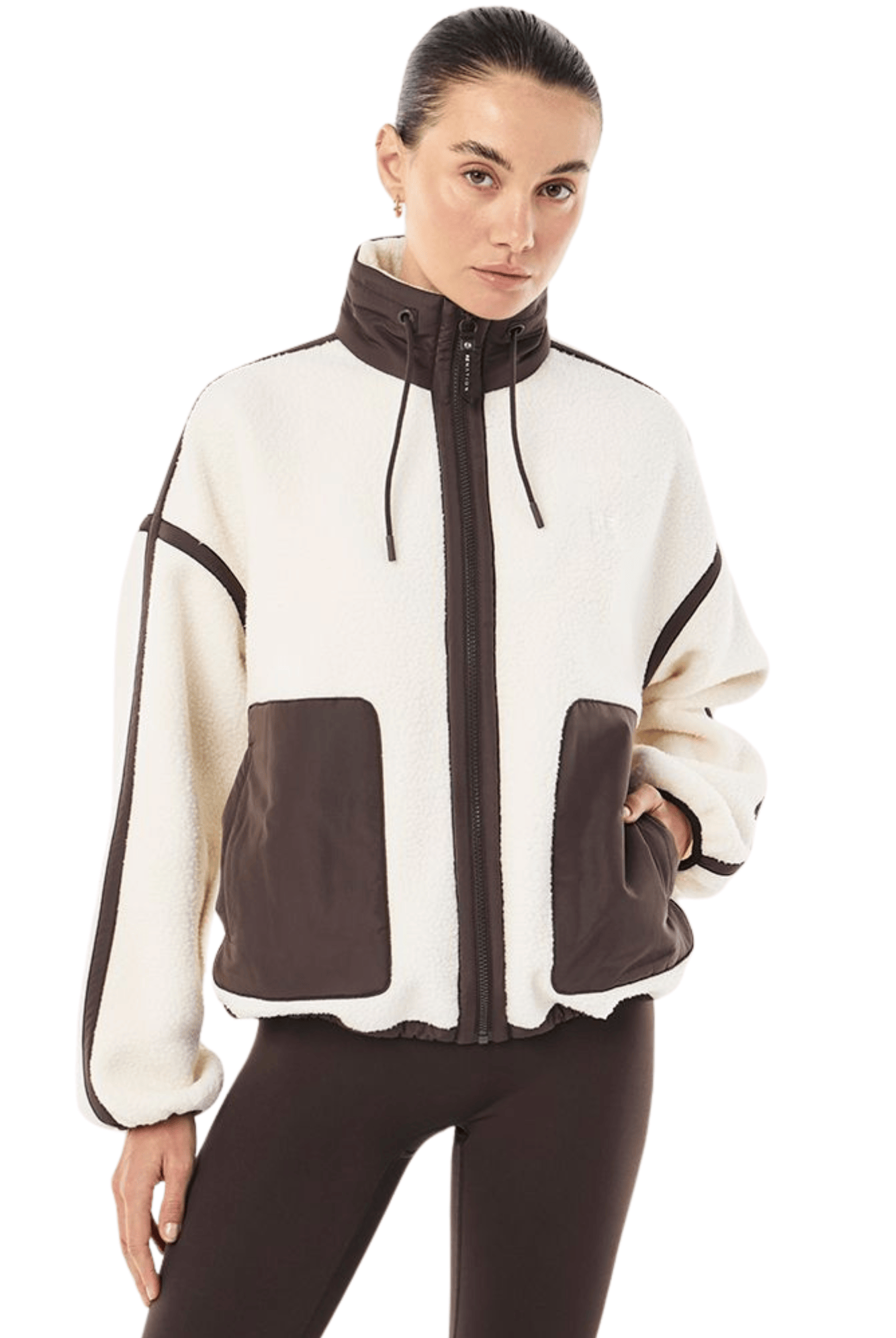 A woman modeling a cropped zip-up jacket with a cream and dark brown color-blocked design, paired with dark brown leggings, against a plain white background.