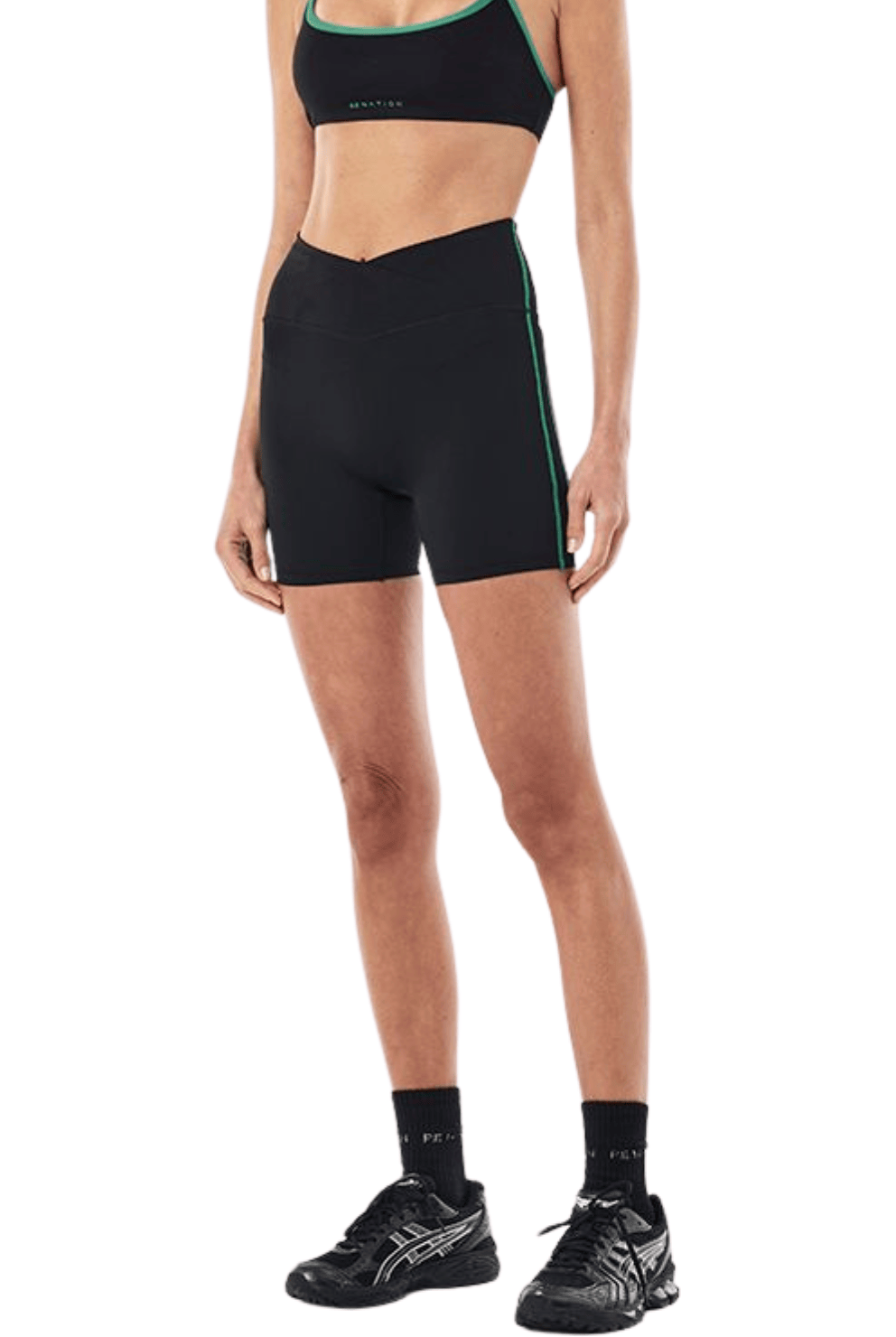A woman modeling a black athletic set consisting of a sports bra and matching biker shorts, both with thin green trim, paired with black socks and running shoes against a plain white background.