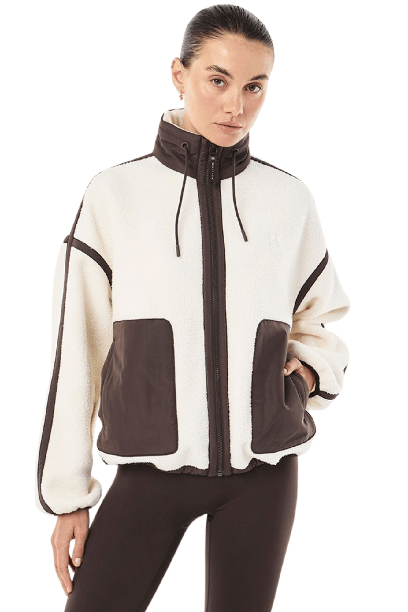 A woman modeling a cropped zip-up jacket with a cream and dark brown color-blocked design, paired with dark brown leggings, against a plain white background.