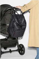 A black quilted backpack with a drawstring closure hanging from the handle of a black stroller, with a person wearing a beige trench coat touching the stroller handle.